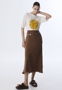 Camiseta blanca con gráfico de sol amarillo, falda marrón de longitud media, cinturón plateado y sandalias marrones con tachuelas y suela gruesa.