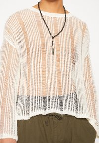 Vit löst stickad långärmad tröja över svart undertröja, olivgröna byxor med dragsko och svart pärlarmband med en fjäderberlock.