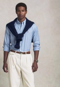 Camisa de botão às riscas azuis e brancas, calças plissadas de cor creme, suéter azul-marinho escuro atado aos ombros e um relógio prateado com mostrador verde.