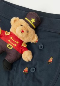 Granatowy materiał z guzikami, ozdobiony małymi motywami misiów. Na materiale spoczywa pluszowy miś ubrany w czerwony mundur i czarny kapelusz.