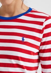 Maglietta a maniche lunghe a righe rosse e bianche con colletto blu e un piccolo logo polo blu sul petto. Materiale in cotone morbido.