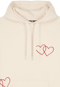 Krēmkrāsas kapucis ar diviem pārklājošiem sarkanu sirds dizainiem apakšējā kreisajā un augšējā labajā pusē. Izgatavots no mīksta auduma ar kapuci.