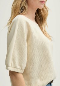 Cremefarbene, strukturierte Bluse mit kurzen Puffärmeln, die ein Karomuster und einen runden Ausschnitt aufweist, kombiniert mit blauen Jeans.