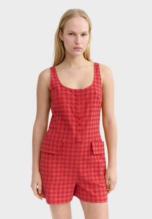 Femme portant une combinaison sans manches rouge à carreaux avec des boutons sur le devant et deux poches avant, debout devant un fond uni.