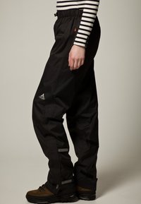 Černé nepromokavé kalhoty s elastickým pasem, reflexními detaily a volným střihem. Natažené přes hnědé turistické boty.