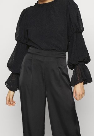 Vrouw draagt een zwarte getextureerde blouse met pofmouwen en hoge taille zwarte satijnen broek tegen een effen achtergrond.