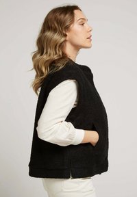 Gilet sans manches noir en tissu texturé, doté d'un col haut et de poches latérales, porté par-dessus une chemise à manches longues blanche.