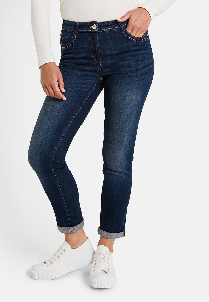 Vaqueros de mezclilla azul oscuro con corte ajustado, puños enrollados, desgaste sutil y costuras en contraste. Combinados con zapatillas blancas.