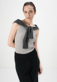 Canotta grigia a coste abbinata a pantaloni neri texturizzati; un maglione grigio scuro legato attorno alle spalle aggiunge un tocco di stile all'outfit.