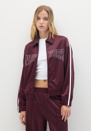 Giacca a vento in satin bordeaux con strisce bianche, con "NEW YORK" stampato in modo prominente sul davanti. Polsini e vita elasticizzati.