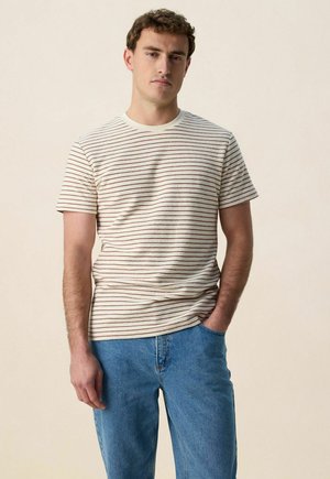 Jeune homme portant un t-shirt crème à fines rayures horizontales marron, assorti à un jean bleu, debout devant un fond uni.