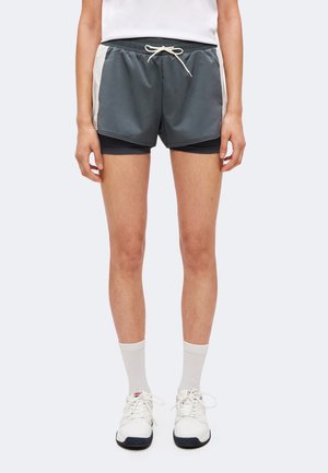 Persona che indossa pantaloncini sportivi grigi a più strati, calzini bianchi alla caviglia, scarpe da ginnastica bianche e una canotta bianca, in piedi davanti a uno sfondo neutro.