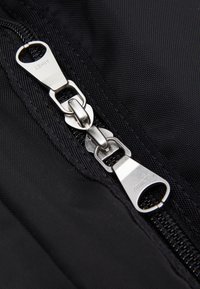 Fermeture éclair en tissu noir avec quincaillerie métallique argentée ; dotée de curseurs doubles et d'un nom de marque en relief sur l'onglet de tirage. Fond texturé.