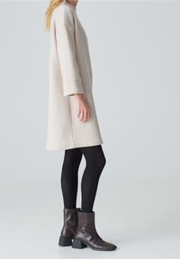 Robe beige clair à manches longues, coupe décontractée, associée à des collants noirs et des bottines marron à talon bloc. Texture lisse.