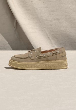Beige suede casual loafer with thick textured sole and moccasin stitching on a neutral background with fabric draped behind.