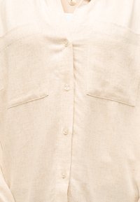 Hele beež linane särk ees olevate nööpide, kahe rinnatasku ja all nähtava valge toppiga.