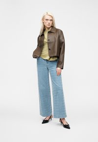 Blonde woman wears brown leather jacket, olive green top, wide-leg light blue patterned jeans, and black kitten heels standing on white background.