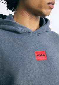 Mikina s kapucňou v sivej farbe, s červeným obdĺžnikovým štítkom s nápisom "HUGO" v čiernej. Mäkká Textúra, jednoduchý dizajn.