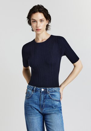 Una donna con capelli corti e scuri indossa un top corto a coste blu navy infilato dentro un paio di jeans blu a vita alta, in piedi con le mani dietro la schiena.