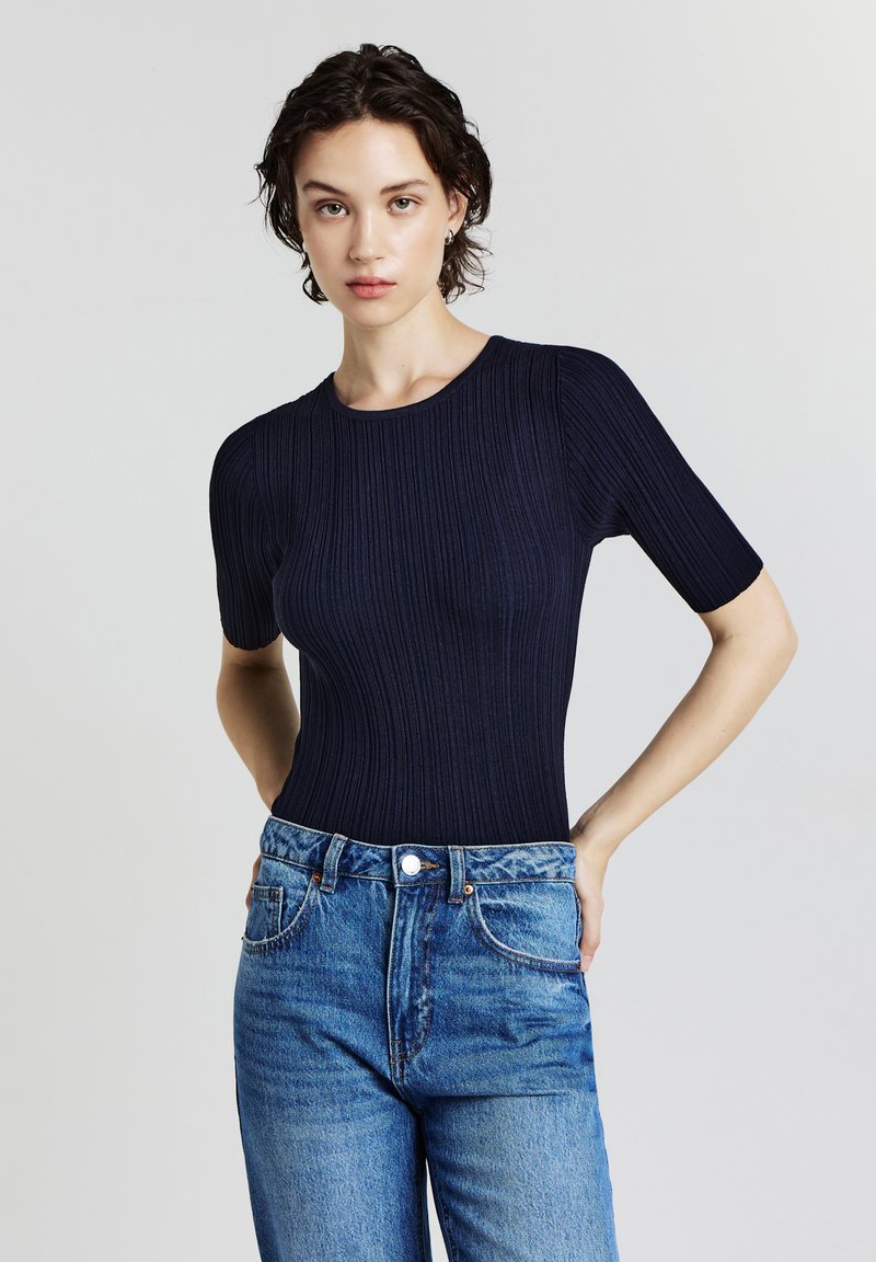 Una donna con capelli corti e scuri indossa un top corto a coste blu navy infilato dentro un paio di jeans blu a vita alta, in piedi con le mani dietro la schiena.