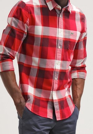 Mann trägt ein rot-weiß-grau kariertes Langarmhemd mit hochgekrempelten Ärmeln und marineblaue Hose, Hände in den Taschen.