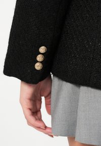 Črn blazer z teksturo, ki ima tri zlate okrogle gumbe na manšeti. Siva krilo je delno vidno pod jakno.