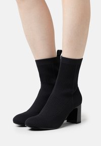 Schwarze, gerippte Knöchelstiefel aus elastischem Material, mit einem klobigen, quadratischen Absatz und einer Zuglasche an der Rückseite für einfaches An- und Ausziehen.