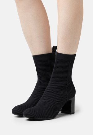 Black ribbed ankle boots made of stretchy material, featuring a chunky square heel and a pull tab at the back for easy wear.