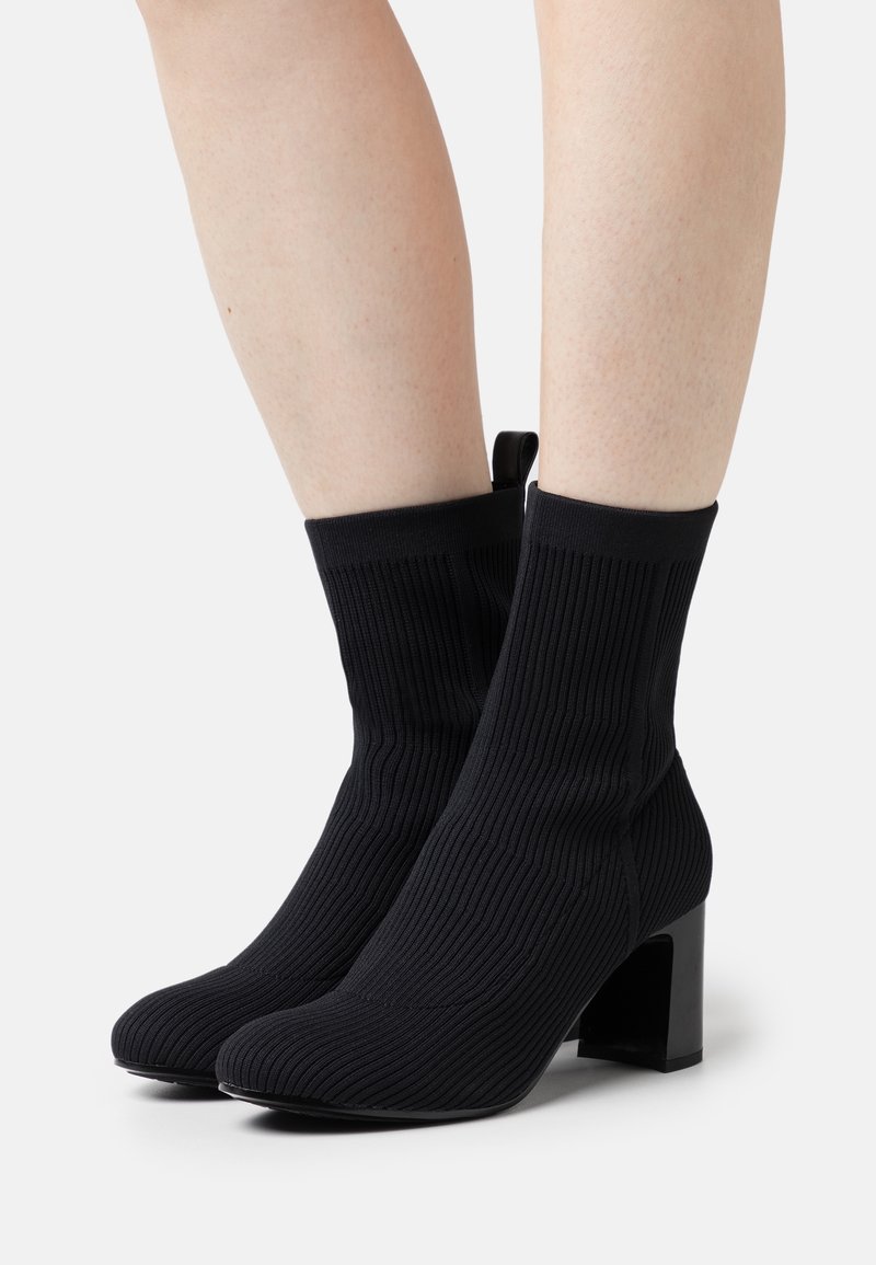 Schwarze, gerippte Knöchelstiefel aus elastischem Material, mit einem klobigen, quadratischen Absatz und einer Zuglasche an der Rückseite für einfaches An- und Ausziehen.