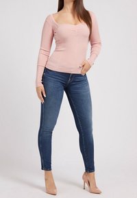 Světle růžové žebrované triko s dlouhým rukávem a hranatým výstřihem, doplněné tmavě modrými úzkými džínami. Modelka má na sobě béžové vysoké podpatky.