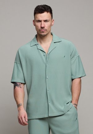 Man met kort donker haar en tatoeages, gekleed in een lichtgroen geribd kort mouwig overhemd met knopen en bijpassende broek, tegen een effen achtergrond.