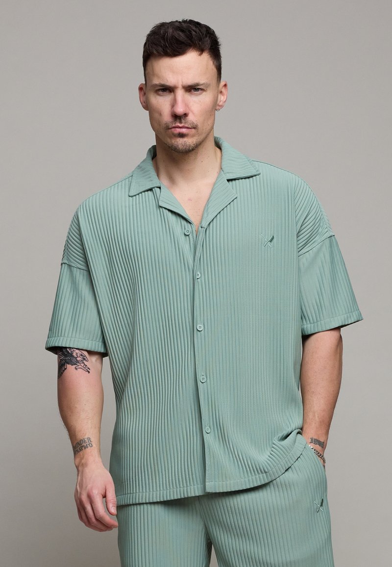 Homme aux cheveux courts et foncés avec des tatouages, portant une chemise à manches courtes côtelée boutonnée vert clair et un pantalon assorti, sur un fond uni.