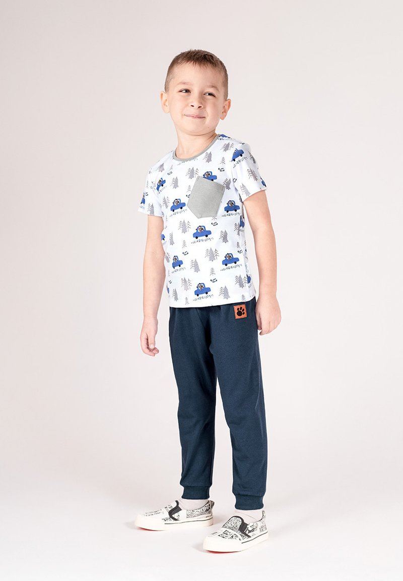 Biały t-shirt z krótkim rękawem z niebieskimi wzorami samochodu i drzewa, szara kieszeń. Ciemnogranatowe spodnie z pomarańczową łatą w kształcie łapy. Jasne buty z graficznym wzorem.