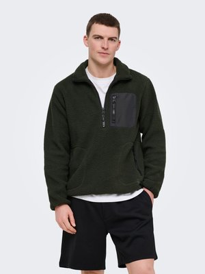 Junger Mann trägt einen dunkelgrünen Fleecepullover mit Reißverschlusstaschen und schwarzen Shorts vor einem schlichten hellen Hintergrund.