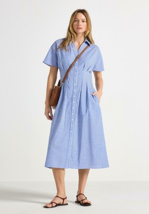 Kvinde iført en blå og hvid ternet midi-kjole med korte ærmer, brun skuldertaske og brune sandaler med stropper.