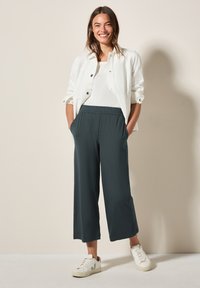 Chemise blanche à manches longues, débardeur blanc, pantalons larges vert foncé, et baskets blanches. Tenue décontractée avec une coupe confortable et des poches latérales.
