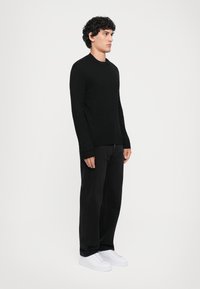 Schwarzer Strickpullover mit rundem Ausschnitt, kombiniert mit schwarzen Hosen und weißen Sneakers, präsentiert an einem Modell, das seitlich steht.