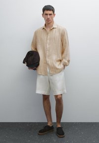 Light beige linen shirt with vertical stripes, white shorts, and black suede shoes. The model holds a dark cap and stands against a neutral background.