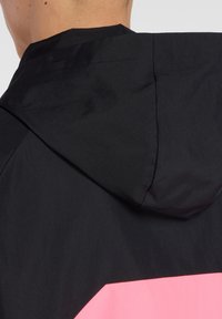 Veste à capuche noire avec un design géométrique rose contrastant dans le dos. Texture lisse, matériau léger, avec des détails de couture visibles.