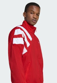 Červená atletická bunda so bielymi pruhmi na ramenách, vysokým golierom, ľahkým materiálom, zipsovým zapínaním a bočnými vreckami.