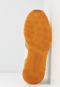 Kengät, joiden pohja on teksturoitua oranssia kumia, vaaleanbeige kangasosa ylöspäin, nojaamassa pystysuoraan valkoisia kulman seiniä vasten.