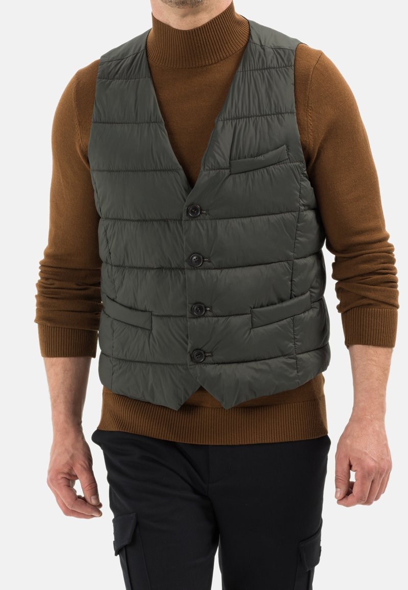 Homme portant un pull à col roulé marron sous un gilet matelassé gris foncé boutonné et un pantalon noir avec poches latérales.