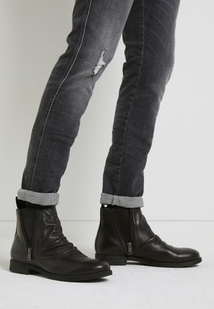 Person trägt schwarze Leder-Stiefeletten mit seitlichen Reißverschlüssen und hochgekrempelte dunkelgraue Jeans mit einem kleinen Riss am rechten Bein.