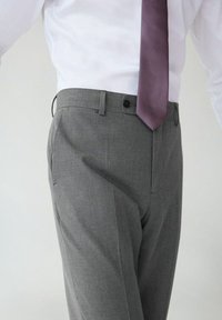 Grijze op maat gemaakte broek van textuurstof, met een platte voorkant, riemlussen en een enkele knoopsluiting. Gecombineerd met een gemusterde stropdas.