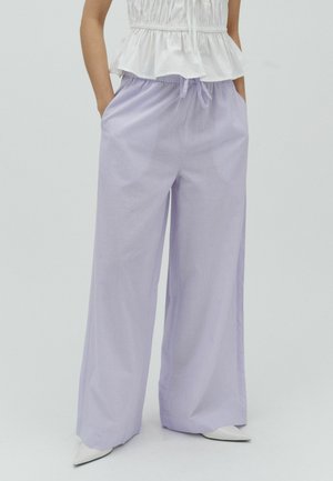 Femme portant un pantalon large ample de couleur violet clair, doté d’une taille élastique et d’un cordon, associé à un top blanc sans manches à volants et des chaussures blanches pointues.