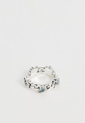 Bague en argent au motif entrelacé et pointu, ornée d’une pierre précieuse bleu pâle au centre, sur fond blanc.