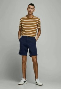 T-shirt rayé marron et crème associé à un short bleu marine, avec un ourlet roulé. Modèle portant des baskets blanches, se tenant devant un fond uni.