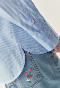 Lichtblauw gestreefd overhemd met afgeronde zoom en knoopmanchetten, gecombineerd met denim shorts met geborduurde bloemen op de achterzak.