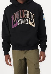 Schwarzer Kapuzenpullover mit mehrfarbig besticktem Text "LOWLIGHTS STUDIOS" in verschiedenen Schriftarten und einem kleinen blauen Etikett am Ärmel.