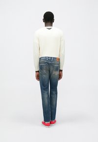 Φόρεμα μακρυμάνικο σε ανοιχτό μπεζ με ριγωτή υφή, συνοδευόμενο από σκούρα μπλε τζιν παντελόνι με ίσια γραμμή και κόκκινα αθλητικά παπούτσια. Το τζιν έχει τσέπες στο πίσω μέρος και ταμπέλα από ταμπά δέρμα.